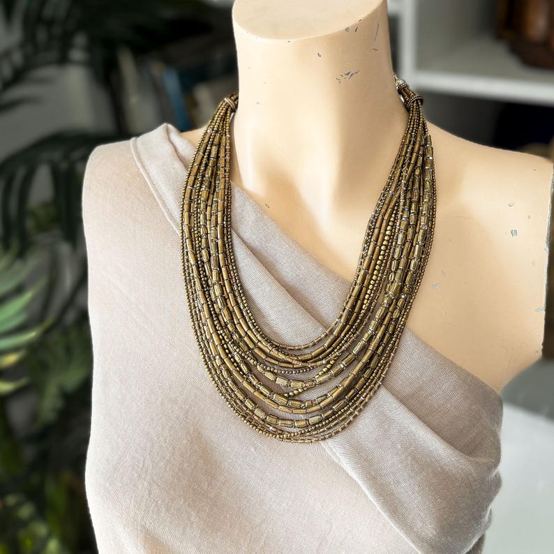 May include: A multi-strand necklace featuring numerous layers of metallic bronze-toned cylindrical and round beads. The necklace is arranged in a cascading, graduated design that rests against the collarbone, creating a textured, layered appearance.