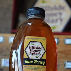 May include: A clear glass bottle of raw honey with a yellow and black label that reads "Hillside Honey Apiary". The label also includes the text "Sweet to the Soul, Health to the Bones." and "Proverbs 16:24". The bottle is sitting on a wooden surface.