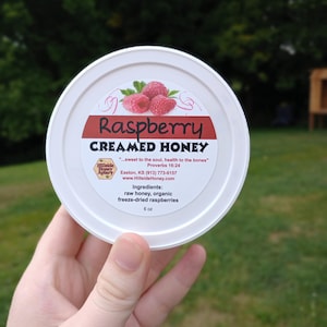 May include: A white container of raspberry creamed honey. The label features a raspberry illustration and the text "Raspberry Creamed Honey." Ingredients include raw honey and freeze-dried raspberries. The container is held in a hand and is 6 oz.