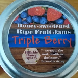 May include: A silver jar with a blue and white label that reads "Honey-sweetened Ripe Fruit Jams Triple Berry" with a quote from Proverbs 16:24. The label also features a hexagon with the words "Hillside Honey Apiary".