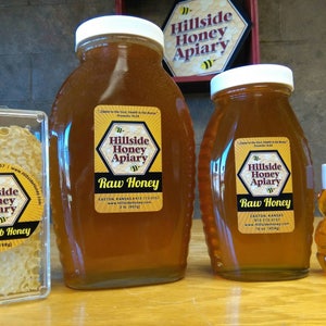 May include: Three glass jars of raw honey from Hillside Honey Apiary in Easton, Kansas. The jars are labeled with the company name and logo. The largest jar is 32 oz, the middle jar is 16 oz, and the smallest jar is 2 oz. There is also a small container of raw comb honey.