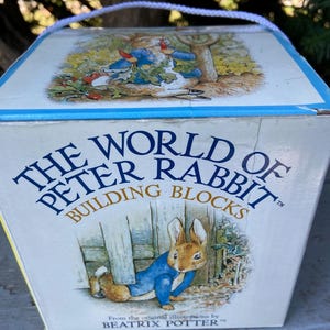 May include: A square cardboard box with a white rope handle featuring The World of Peter Rabbit Building Blocks text. The box displays illustrations of Peter Rabbit in a blue jacket, with the text From the original illustrations by Beatrix Potter.
