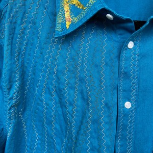 Puede incluir: Una camisa azul turquesa con botones y un cuello bordado en amarillo y dorado. La camisa tiene una etiqueta que dice "100% algod&oacute;n" y "LAND".