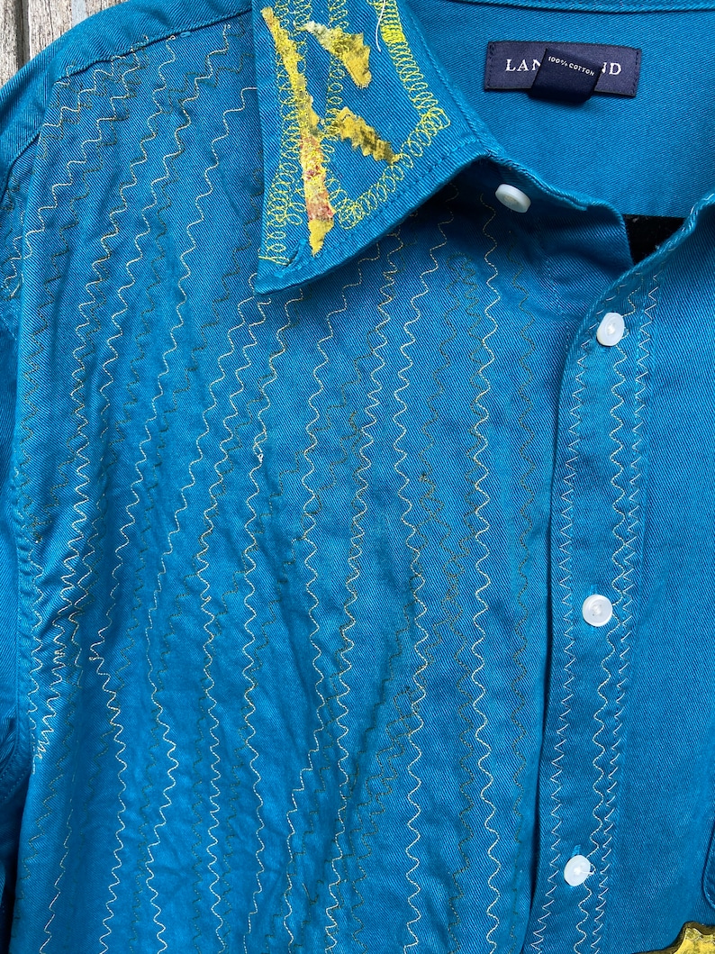 Puede incluir: Una camisa azul turquesa con botones y un cuello bordado en amarillo y dorado. La camisa tiene una etiqueta que dice "100% algod&oacute;n" y "LAND".