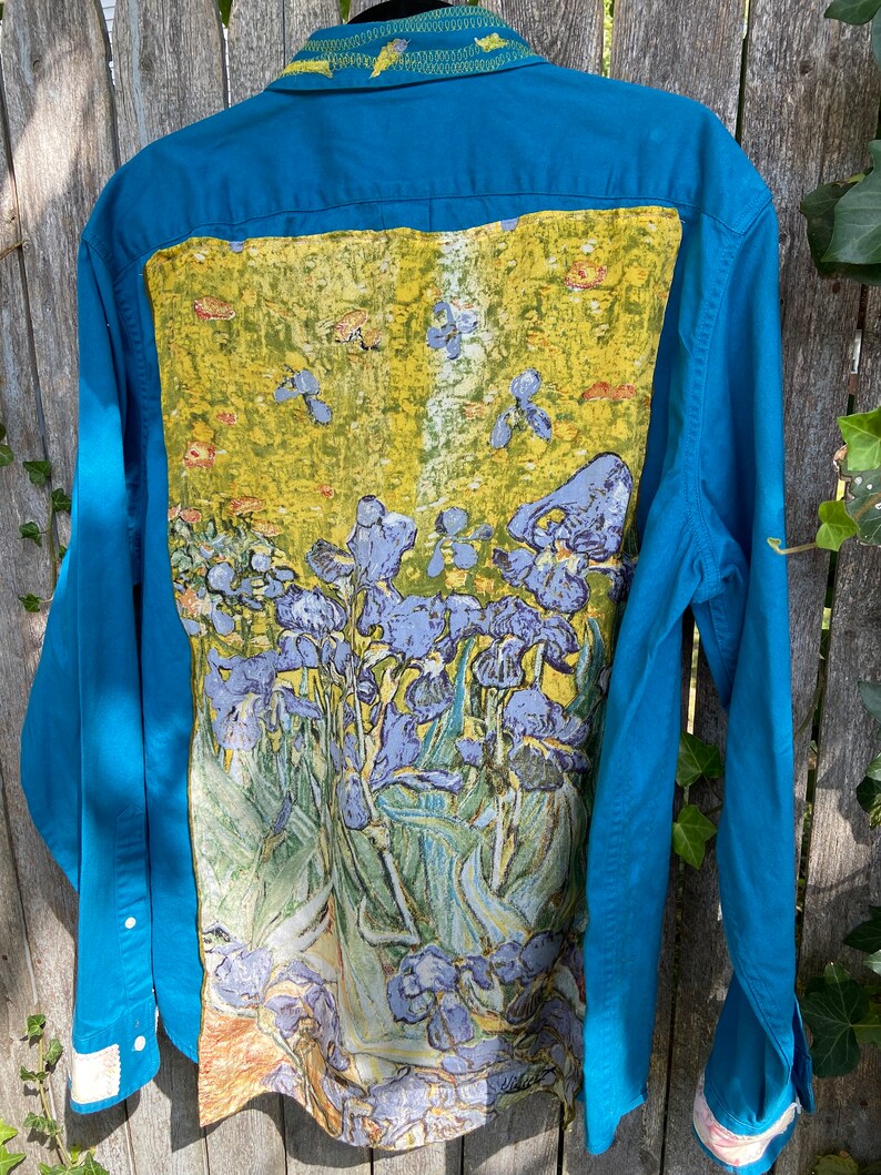 Puede incluir: Una camisa de manga larga azul turquesa con botones con una pintura de iris morados en la espalda. La pintura est&aacute; sobre un fondo amarillo con hojas verdes.