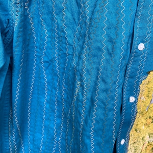 Puede incluir: Una camisa azul turquesa con un patr&oacute;n de puntadas en zigzag decorativo. La camisa tiene un frente abotonado y mangas largas.