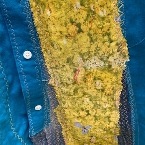 Puede incluir: Una camisa azul turquesa con un tejido estampado floral amarillo y verde cosido en la parte delantera. El tejido est&aacute; cosido con una puntada en zigzag dorada.