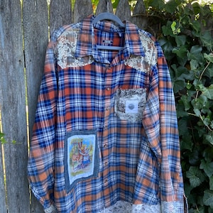 May include: A plaid button-down shirt in blue, orange, and white, with lace accents on the shoulders, cuffs, and hem. A patch with a vintage illustration is sewn on the front. The shirt is hanging on a wooden fence.