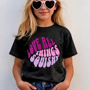 Könnte beinhalten: Ein dunkelgraues T-Shirt mit dem Text "LOVE ALL THINGS SQUISHY" in einem rosa und lila Design. Die Person auf dem Bild trägt herzförmige Sonnenbrillen und blaue Jeans.