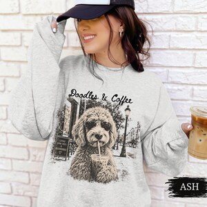 May include: An ash gray crewneck sweatshirt featuring a black and white graphic print of a doodle dog wearing sunglasses and holding a coffee cup. The text Doodles & Coffee is printed above the dog, with a street scene background including a lamp post.
