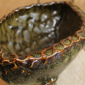 May include: A brown and green ceramic planter with a textured, uneven surface and a scalloped rim. The planter has a speckled glaze and a glossy finish.