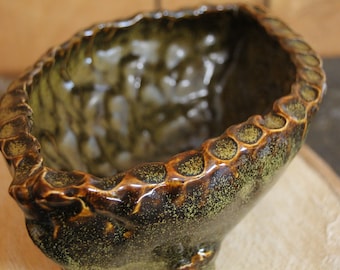 May include: A brown and green ceramic planter with a textured, uneven surface and a scalloped rim. The planter has a speckled glaze and a glossy finish.