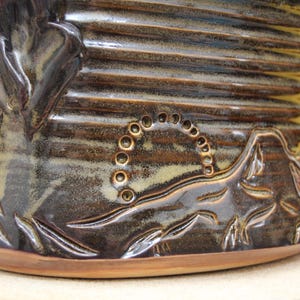 May include: A brown ceramic vase with a textured surface and a raised design of leaves and a circle of dots. The vase has a wide base and a narrow neck.