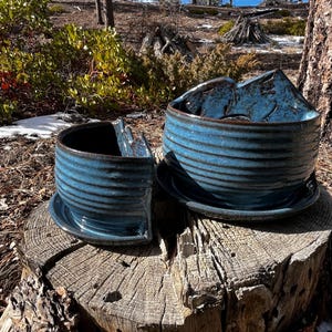 May include: Two handmade ceramic bowls with matching saucers. The bowls are a vibrant blue with a textured, ribbed design and a dark brown rim. They are placed on a weathered tree stump, with a natural outdoor background.