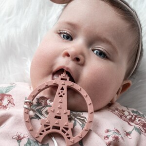 K&ouml;nnte beinhalten: Ein Baby in einem rosa, blumigen Strampler mit einem rosa Schleifen-Haarband kaut auf einem rosa Silikon-Bei&szlig;ring in Form des Eiffelturms.