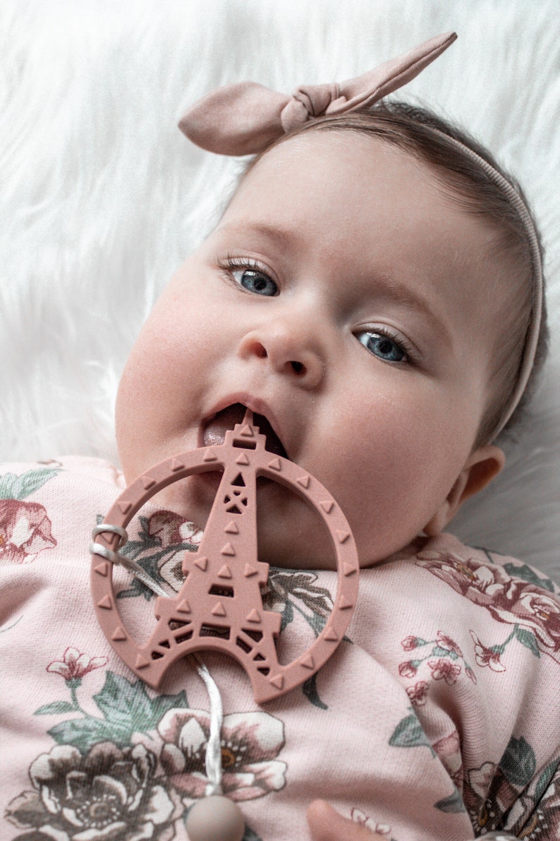 K&ouml;nnte beinhalten: Ein Baby in einem rosa, blumigen Strampler mit einem rosa Schleifen-Haarband kaut auf einem rosa Silikon-Bei&szlig;ring in Form des Eiffelturms.