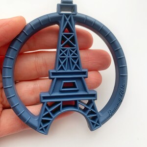 K&ouml;nnte beinhalten: Ein blauer Silikon-Bei&szlig;ring in Form des Eiffelturms mit einem Kreis darum. Der Bei&szlig;ring hat den Text "Gummi Chic" an der Seite.