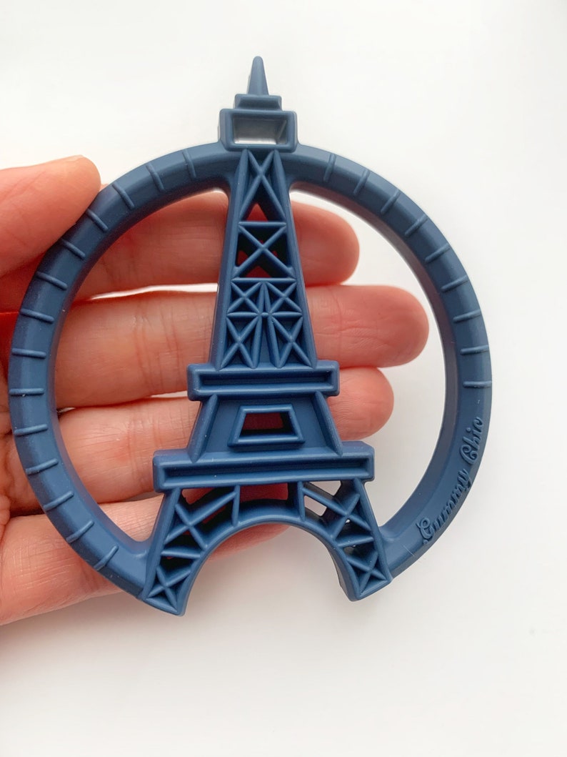K&ouml;nnte beinhalten: Ein blauer Silikon-Bei&szlig;ring in Form des Eiffelturms mit einem Kreis darum. Der Bei&szlig;ring hat den Text "Gummi Chic" an der Seite.