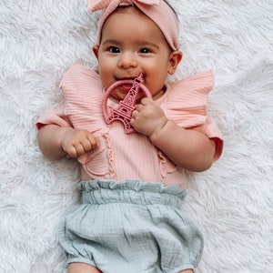 K&ouml;nnte beinhalten: Ein Baby tr&auml;gt einen rosa R&uuml;schen-Body und eine hellblaue R&uuml;schenwindel, mit einem rosa Schleifen-Stirnband, h&auml;lt einen rosa Eiffelturm-Bei&szlig;ring.