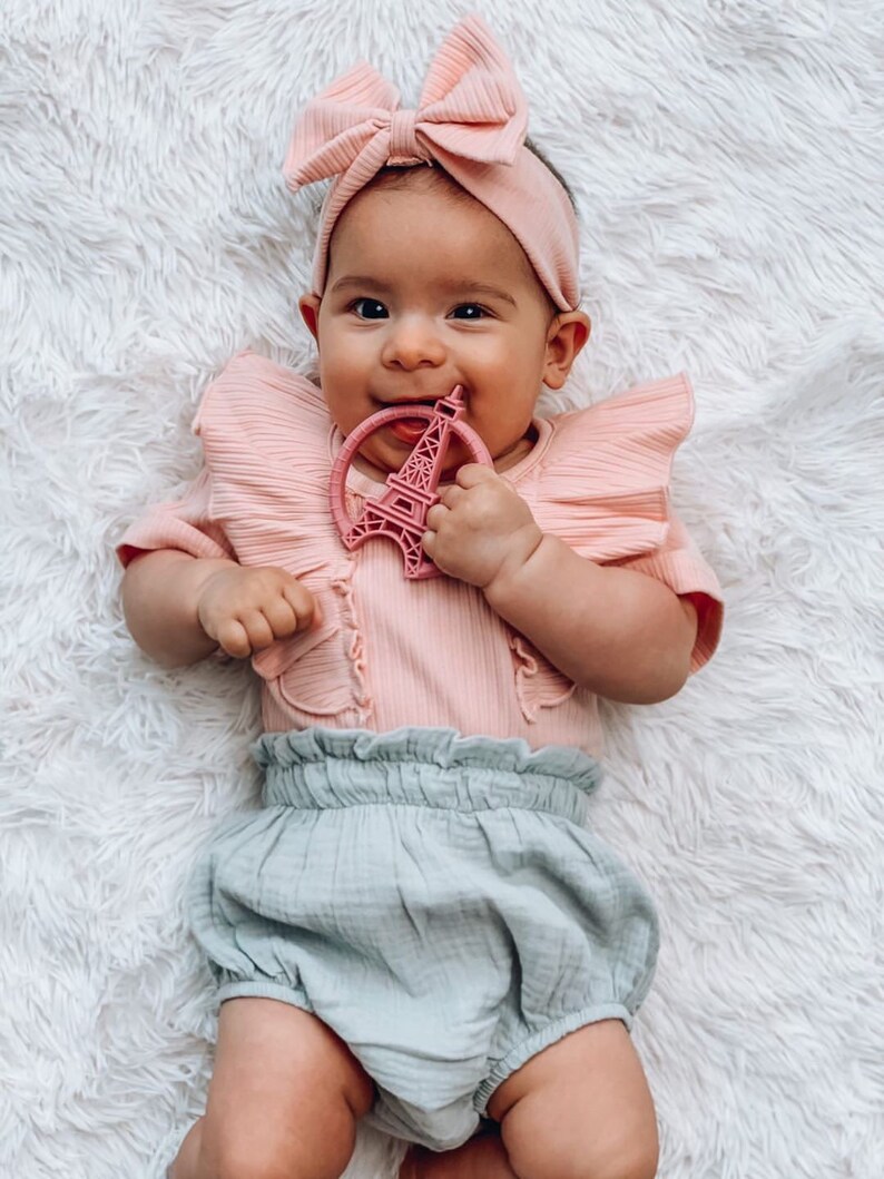 K&ouml;nnte beinhalten: Ein Baby tr&auml;gt einen rosa R&uuml;schen-Body und eine hellblaue R&uuml;schenwindel, mit einem rosa Schleifen-Stirnband, h&auml;lt einen rosa Eiffelturm-Bei&szlig;ring.