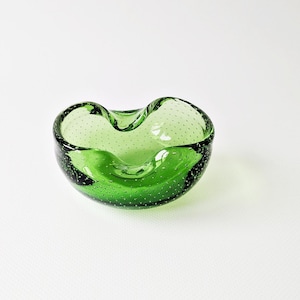 May include: A small, green glass ashtray with a bubbly texture. The ashtray has a unique, curved shape with a smooth, glossy finish. The color is a vibrant, transparent green, and the overall design is simple and elegant.