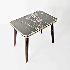 Peut inclure: Petite table d'appoint rectangulaire avec un plateau noir effet marbre et des pieds effilés brun foncé. Le plateau est bordé d'une garniture dorée. La table mesure environ 61 cm de long, 41 cm de large et 51 cm de haut.