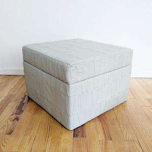 Może przedstawiać: Biały skórzany puf z kwadratowym blatem i wzorem tkanym. Puf stoi na drewnianej podłodze.