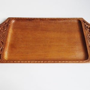 Könnte beinhalten: Rechteckiges Holztablett mit geschnitzten Blumen- und Rankenmustern an den Enden. Das Tablett ist aus hellbraunem Holz gefertigt und hat eine glatte, ebene Oberfläche. Die Kanten sind leicht erhöht und haben eine dekorative Bordüre.