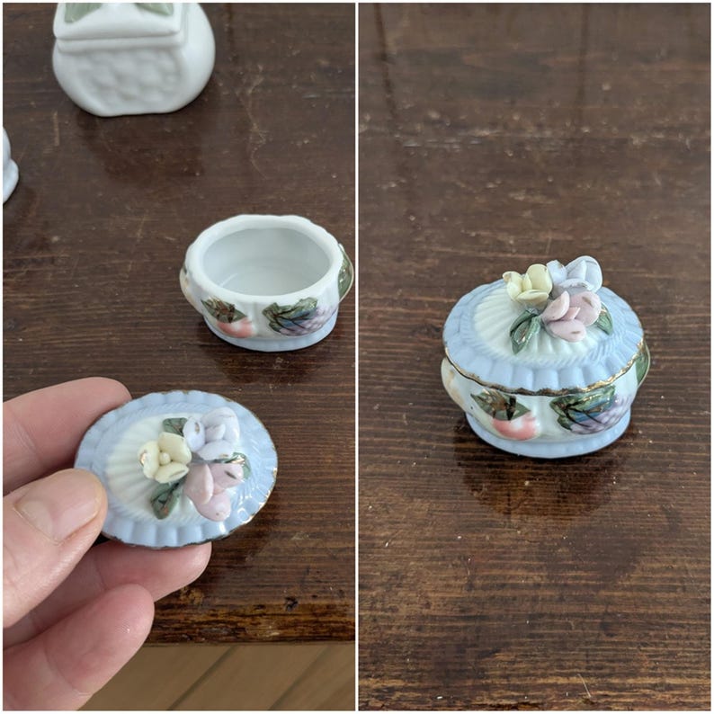May include: Three small porcelain trinket boxes with floral designs. The boxes are white with blue and gold accents. One box has a lid with a raised floral design. The other two boxes have lids with a raised floral design and a raised fruit design.