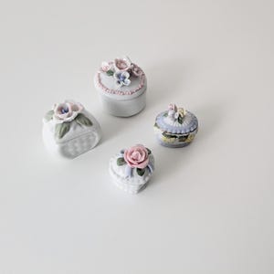 May include: Four small porcelain trinket boxes with floral designs. The boxes are white with pastel pink, blue, and green accents. Each box has a different floral design with a single rose or a cluster of flowers.