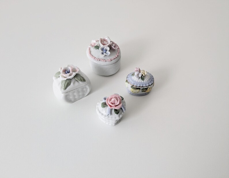 May include: Four small porcelain trinket boxes with floral designs. The boxes are white with pastel pink, blue, and green accents. Each box has a different floral design with a single rose or a cluster of flowers.