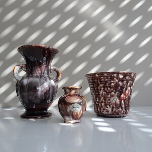 May include: Three ceramic vases in different sizes and styles. The largest vase is a brown and white glazed amphora with two handles. The medium vase is a brown and white glazed cylinder with a label that reads "Kay-Bo Germany". The smallest vase is a brown and white glazed cylinder.