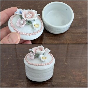 May include: A white porcelain trinket box with a floral design. The lid features pink, white, and blue flowers with green leaves. The box has a white and pink beaded trim.