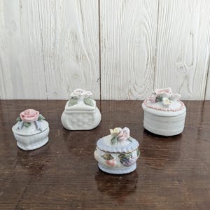 May include: Four small porcelain trinket boxes with floral and fruit designs. The boxes are white with pastel pink, blue, and green accents.