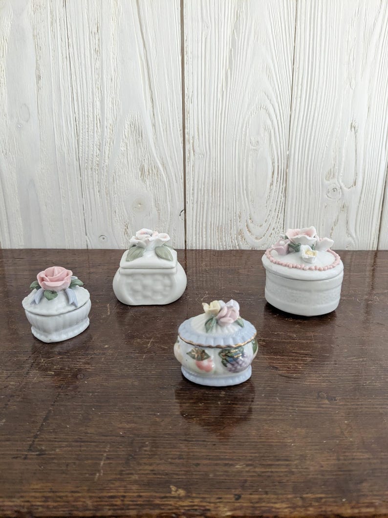 May include: Four small porcelain trinket boxes with floral and fruit designs. The boxes are white with pastel pink, blue, and green accents.