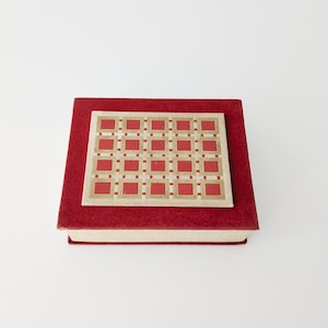 Op de afbeelding: Een rood boek met een decoratief rechthoekig ontwerp op de omslag. Het ontwerp heeft een raster van rode vierkanten en gouden accenten. Het boek staat op een witte achtergrond.