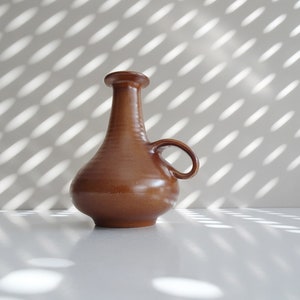 May include: A brown ceramic vase with a single handle. The vase has a rounded body and a narrow neck.