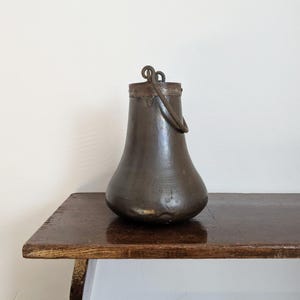 Op de afbeelding: Een donkerbruine metalen kan met een gebogen handvat. De kan heeft een brede basis en een smalle hals. Hij staat op een houten tafel.