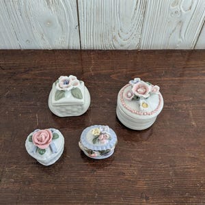 May include: Four small porcelain trinket boxes with floral designs. The boxes are white with pink, blue, and green flowers. The boxes are in the shape of a square, heart, oval, and a rounded rectangle.