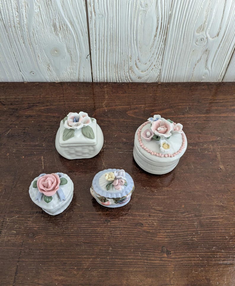 May include: Four small porcelain trinket boxes with floral designs. The boxes are white with pink, blue, and green flowers. The boxes are in the shape of a square, heart, oval, and a rounded rectangle.