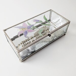 Könnte beinhalten: Eine transparente Glas-Schmuckschatulle mit silbernem Metallrahmen und einem gemalten Blumendesign. Die Schachtel ist geöffnet und enthält eine silberne Kettenkette.
