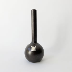 May include: A dark brown ceramic vase with a round base and a long, slender neck. The vase has a matte finish and a small, rectangular label near the base. The vase is likely for decorative purposes.
