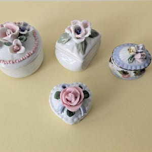 May include: Four small porcelain trinket boxes with floral designs. The boxes are white with pink, blue, and green flowers. One box is heart-shaped, one is oval, and two are square.
