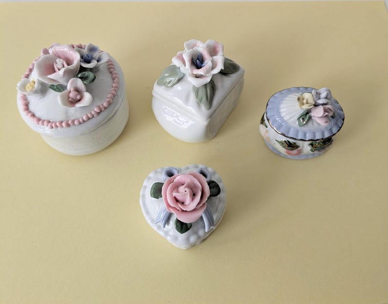 May include: Four small porcelain trinket boxes with floral designs. The boxes are white with pink, blue, and green flowers. One box is heart-shaped, one is oval, and two are square.