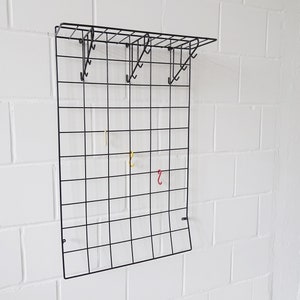 Peut inclure: Organiseur mural en grille métallique noire avec une étagère et des crochets. La grille est en fil métallique et a une finition noire. Il y a plusieurs crochets sur la grille, dont certains sont utilisés pour accrocher des objets colorés.