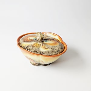 以下が含まれることがあります： ユニークな有機的な形状の、蓋付きのセラミック製のボウル。 蓋とボウルは、茶色、オレンジ、黄色などのアースカラーのブレンドで、テクスチャ加工された表面が特徴です。 ボウルは3つの小さな足の上にあり、蓋には小さなハンドルが付いています。