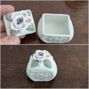 May include: A white porcelain trinket box with a floral design. The lid features a pink and white flower with green leaves. The box has a woven pattern.