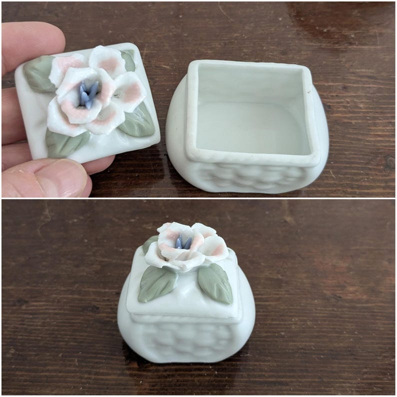 May include: A white porcelain trinket box with a floral design. The lid features a pink and white flower with green leaves. The box has a woven pattern.