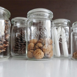 May include: A selection of clear glass jars, each containing different natural items. The jars hold pine cones, walnuts, and white candles. The jars are displayed on a white surface.