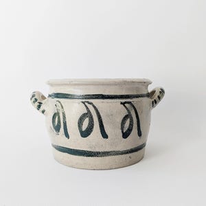 Puede incluir: Una olla de cerámica de color crema con dos asas y detalles pintados en azul oscuro. La olla presenta una banda azul oscuro alrededor de la base y una serie de formas estilizadas en azul oscuro alrededor del cuerpo. La olla es probablemente para uso decorativo o funcional.